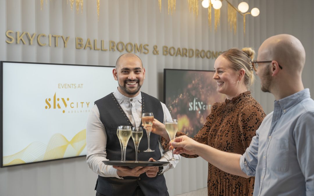 Inside a 5 Star Hotel, a duty manager is carrying a tray of champagne flutes. Two guests approach and take two champagne flutes.