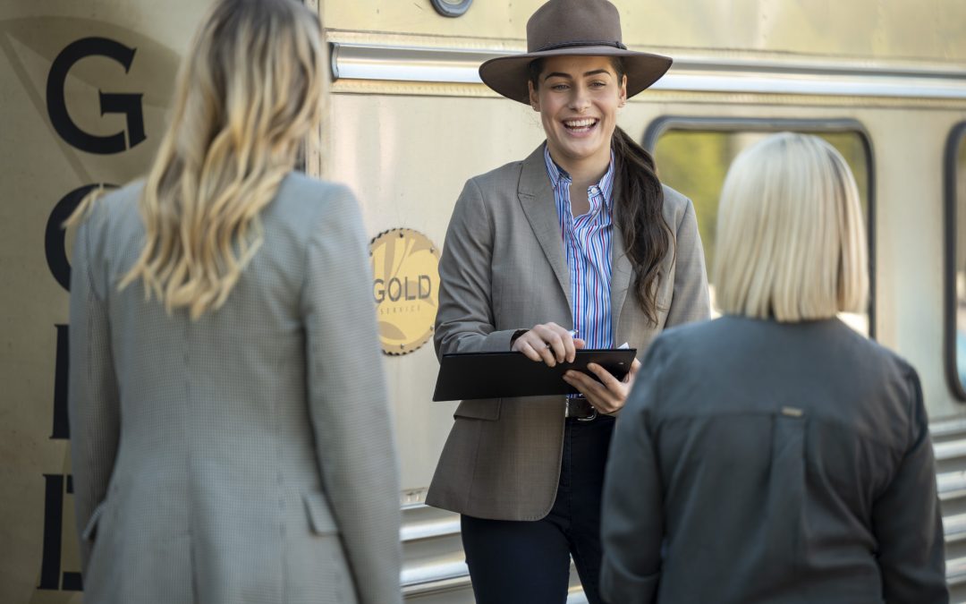 A young woman in a neutral blazer and broad brim hat greets two guests boarding a train.