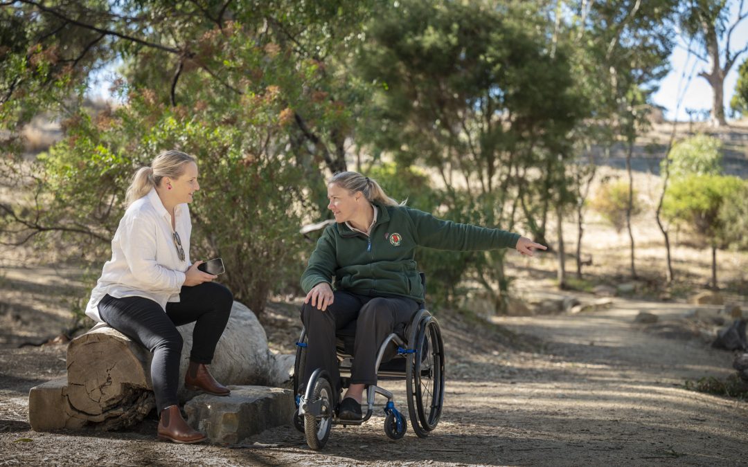 A woman in a wheelchair speaks with another woman seated on a wooden log in Australian bush.